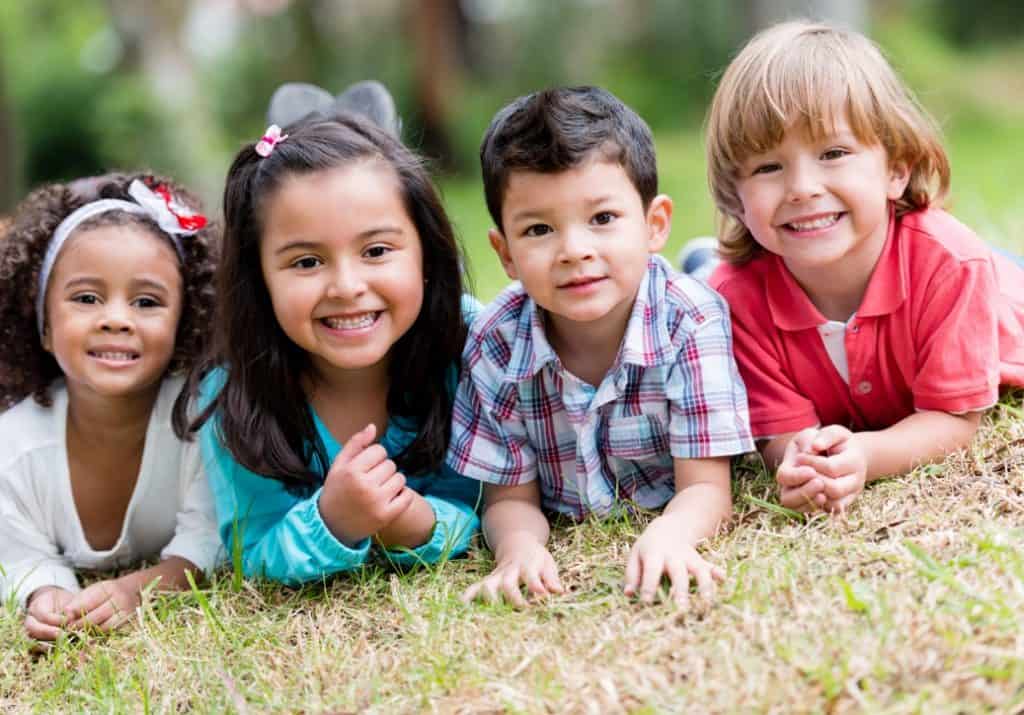 Four toddler age children on their tummies on the grass with their heads up with bigs smiles, two girls to the left, African American and Hispanic, and one boy hispanic and one Caucasian boy. Looking all ready to play