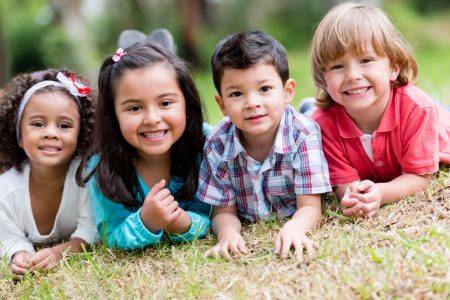 Four toddler age children on their tummies on the grass with their heads up with bigs smiles, two girls to the left, African American and Hispanic, and one boy hispanic and one Caucasian boy. Looking all ready to play